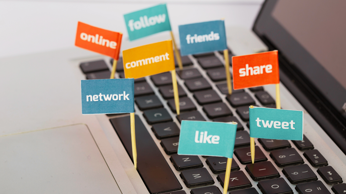 A laptop keyboard with small colorful flags displaying words like "online," "follow," "friends," "share," "comment," "network," "like," and "tweet," symbolizing social media engagement.